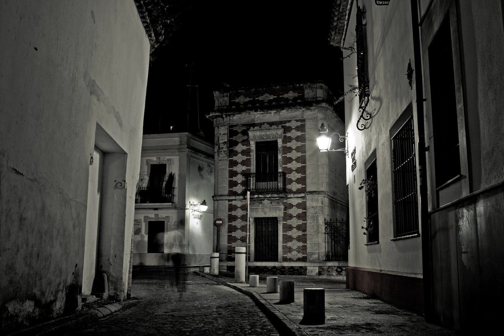 Fotografía en blanco y negro de una calle empedrada y serpenteante en el casco histórico de Córdoba. Un edificio con patrón de ladrillos en damero se destaca entre las fachadas encaladas con faroles, rejas y balcones de hierro forjado. La calle vacía y la iluminación dramática crean una atmósfera misteriosa y evocadora.