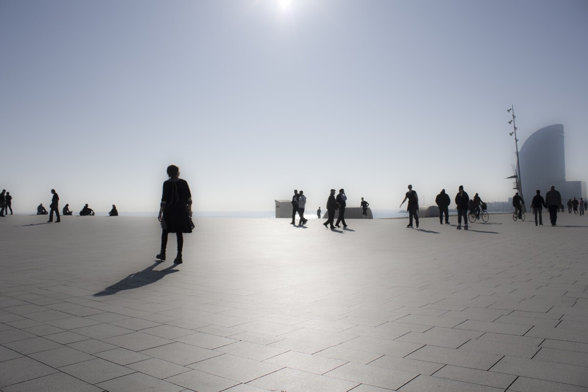 Siluetas de personas caminando y sentadas en un paseo pavimentado en Port Vell, Barcelona, con el mar y el Hotel W visibles en el fondo.